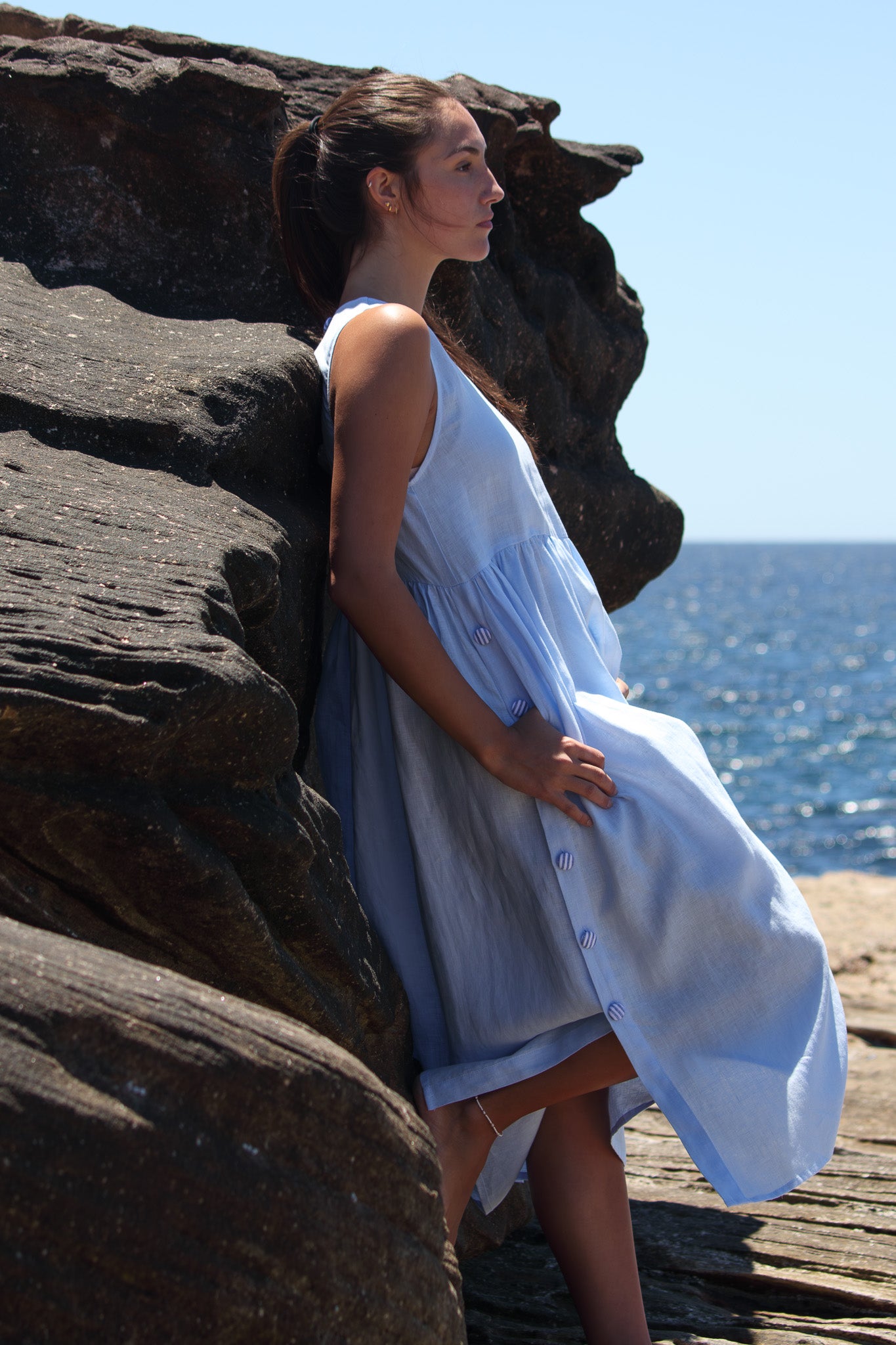 Side profile of the Wonka Dress in sky blue linen, showing the striped covered buttons and soft drape as she leans against the rocks.