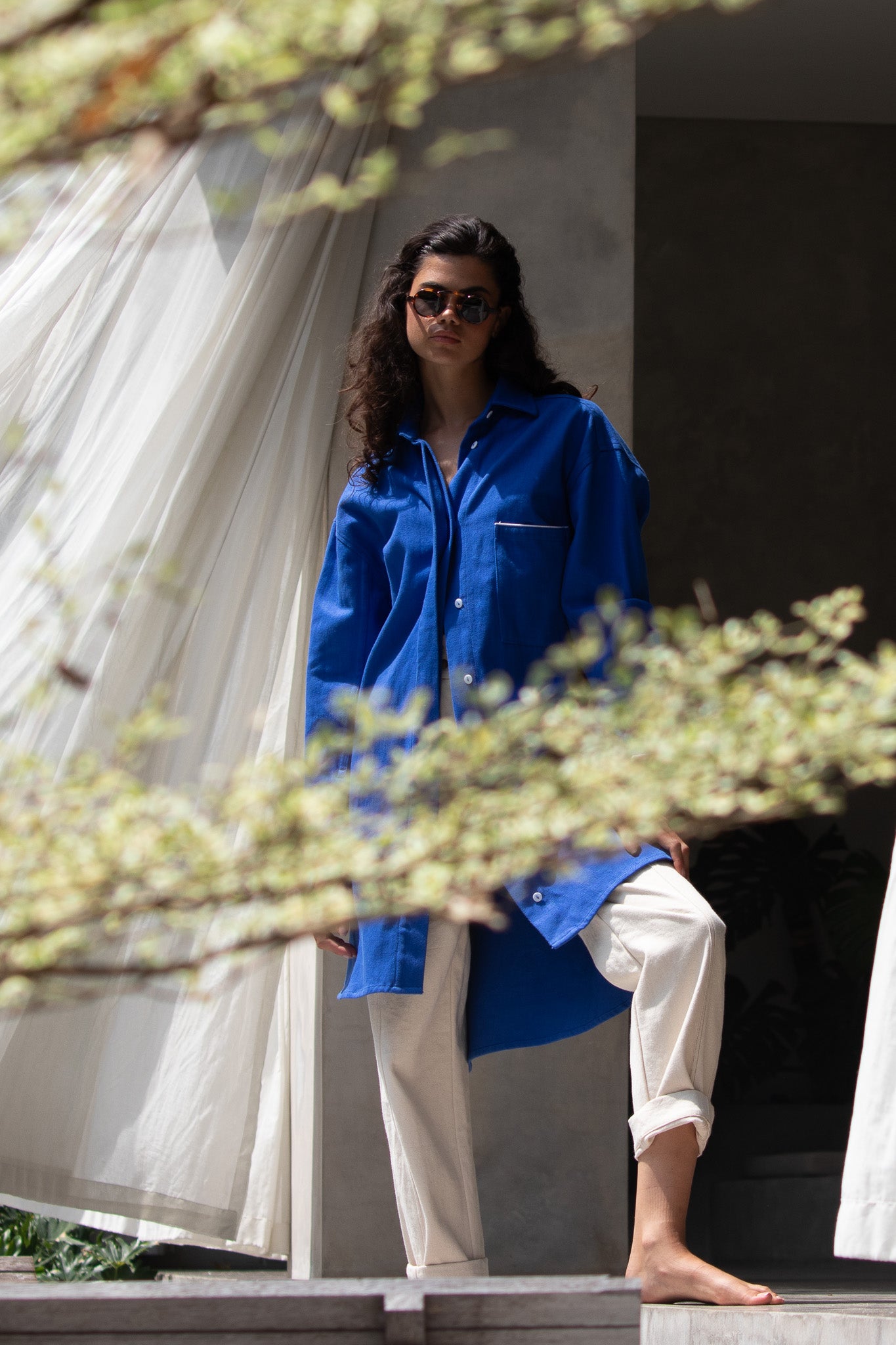 model looking through a gap in a tree standing on her porch of her villa wearing denim blue jacket 
