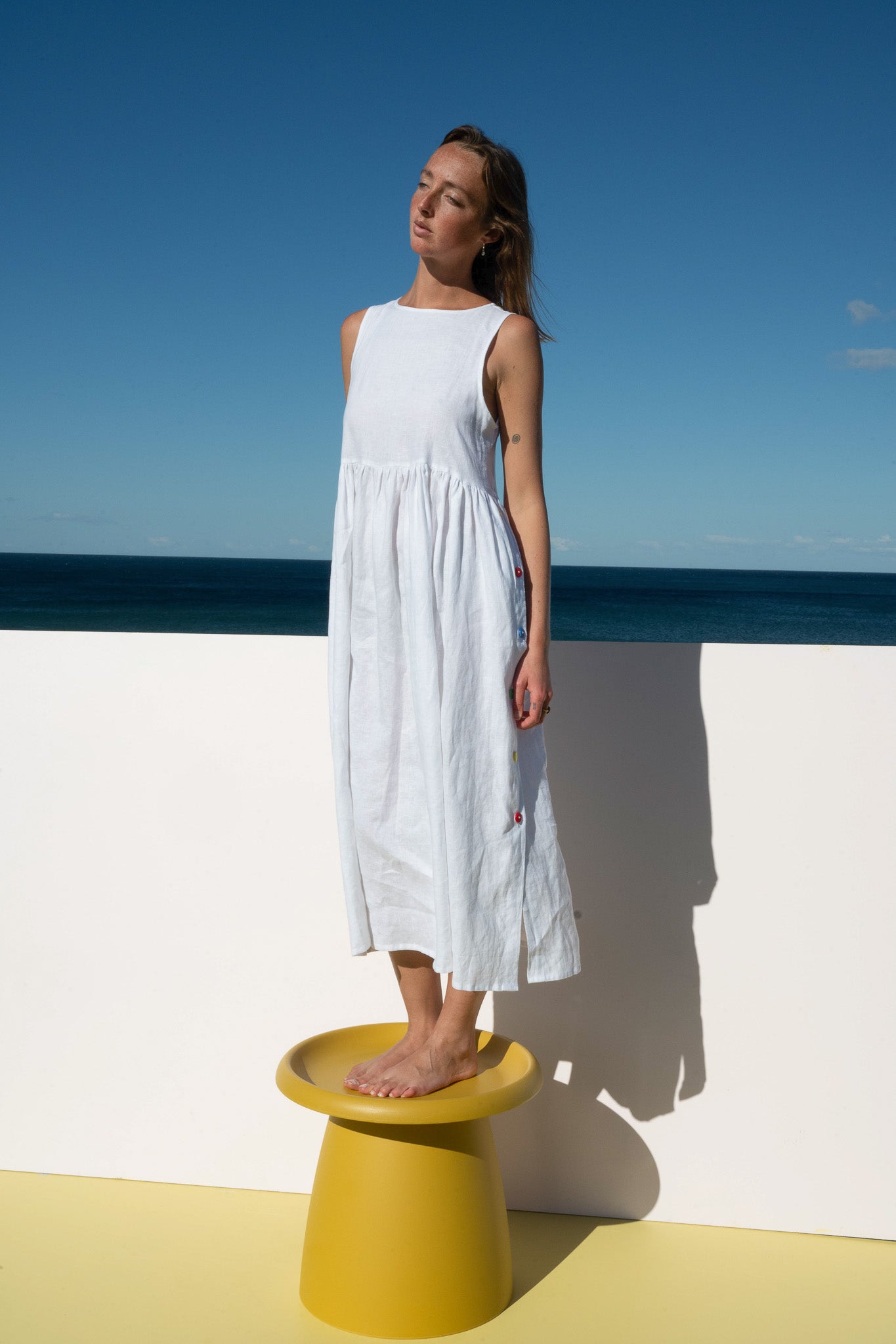 Model wearing the Wonka Sleeveless Dress in linen, standing on a yellow stool with ocean backdrop.