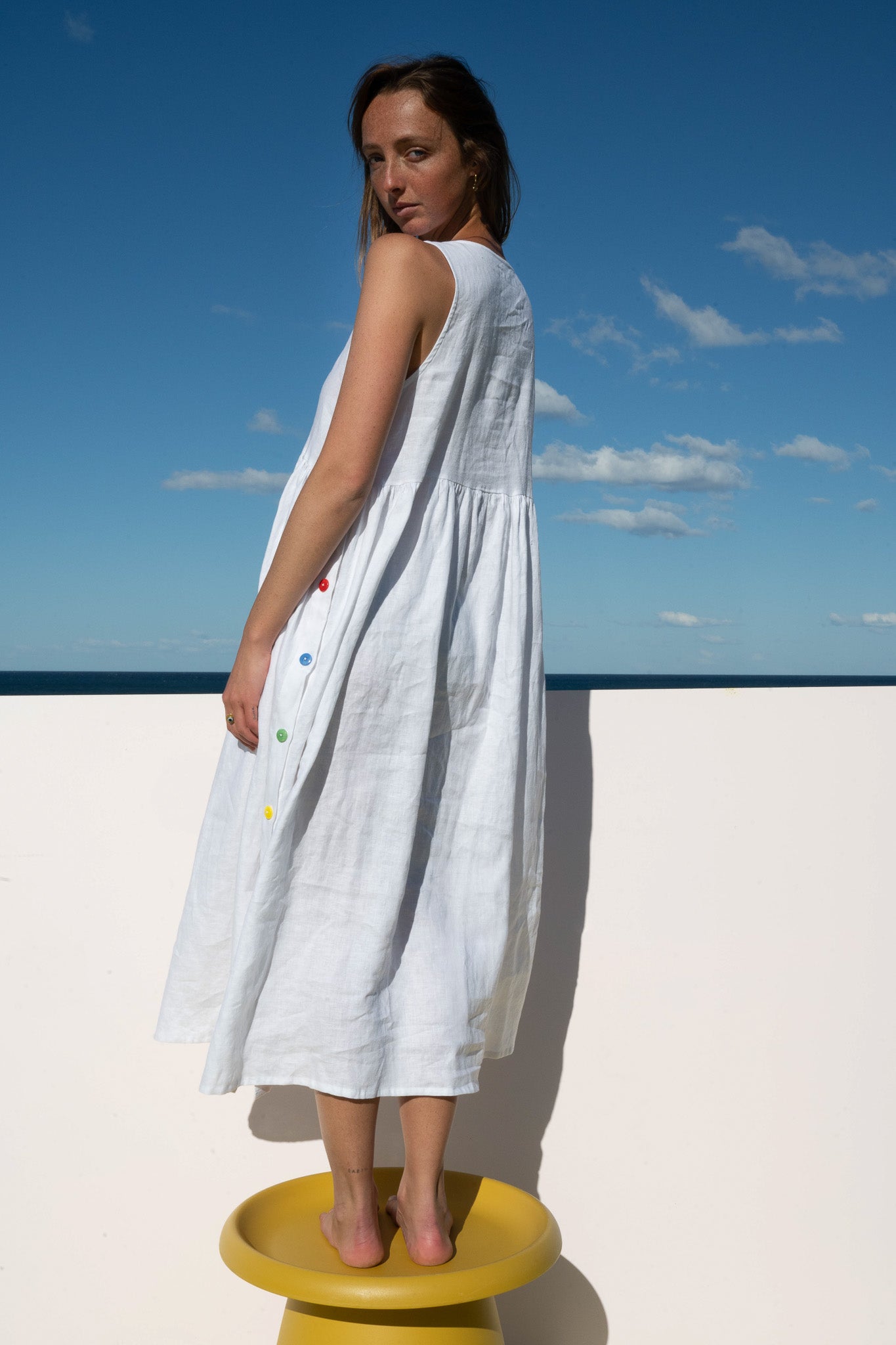 Model wearing the Wonka Sleeveless Dress in white linen with rainbow buttons, back view by the ocean.